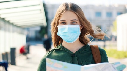 Close up portrait of cheerful Caucasian young woman traveller in medical mask standing outdoor in...