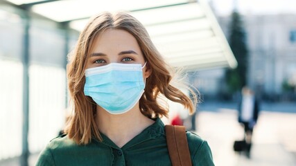 Close up portrait of cheerful Caucasian young woman in medical mask standing outdoor on sunny street on bus stop and looking at camera in good mood. Coronavirus pandemic, covid-19 concept