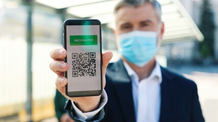 Close up of middle-aged Caucasian man traveller standing outdoor on street in medical mask holding smartphone showing QR code with covid-19 vaccination digital passport, vaccination certificate