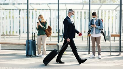 Obraz premium People in medical masks standing on bus stop or train station keeping safe social distance and using devices. Woman traveller with suitcase typing on cellphone, male calling on smartphone outdoors