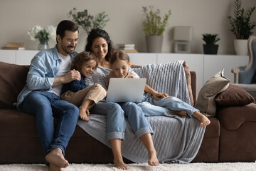Happy multiethnic family with two little kids relax on sofa in living room use laptop together. Smiling multiracial parents with biracial children rest at home talk on video webcam call on computer.