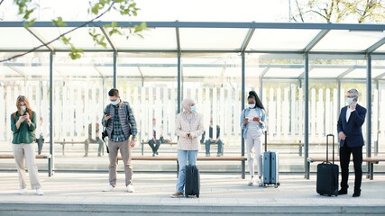 Different mixed-races people with masks on faces at bus stop or train station with suitcases keeping social distance. Man talking on cellphone. Woman texting on smartphone, travellers, Covid concept