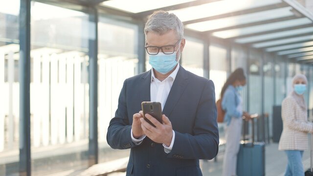 Portrait of handsome adult middle-aged Caucasian man in medical mask standing on street in city and texting on smartphone browsing online. people travellers with suitcases on background