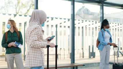 Portrait side view of Muslim woman traveller in medical mask with suitcase walking by bus stop and typing on cellphone gadget. Mixed-race tourists people on background. Covid-19 pandemic travel