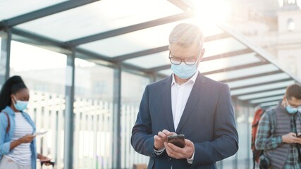 Portrait of Caucasian concentrated senior man traveller in mask standing on street outdoors at bus stop and typing on smartphone browsing online. Mixed-race tourists with suitcases on background