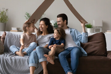 Overjoyed young family with small children relax in new own house excited moving relocating. Happy parents renters hold paper roof relax with kids at home, take bank loan mortgage. Rental concept.