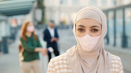 Close up of Muslim young beautiful female wearing mask on face looking at camera while standing in town at bus stop waiting outdoors. People travellers on background, coronavirus pandemic, new reality