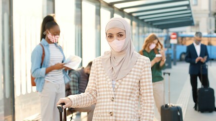 Obraz premium Close up of Muslim young beautiful female wearing mask on face looking at camera while standing in town at bus stop waiting outdoors. People travellers on background, coronavirus pandemic, new reality