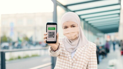 Muslim woman in medical mask showing an International Vaccination Certificate on smartphone. QR...