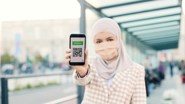 Muslim woman in medical mask showing an International Vaccination Certificate on smartphone. QR code of vaccinated person on cellphone app. Close up concept. Digital health passport