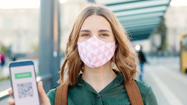 Caucasian happy woman in mask showing an International Vaccination Certificate on smartphone. QR code of vaccinated person on cellphone app. Close up. Digital health vaccine passport