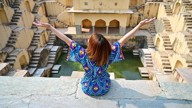 Cheerful woman wearing colorful dress raising arms sitting over Indian old step-well in Rajasthan. slow-motion footage
