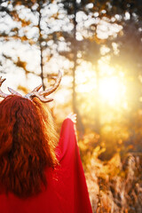 Woman in long red dress with deer horns in autumn forest. A fairy creature wanders in the autumn forest and studies nature.