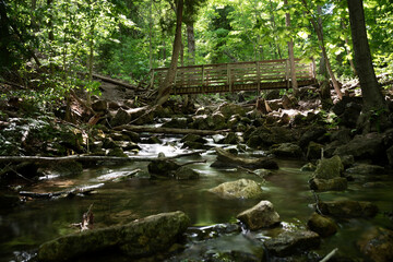 Fototapeta na wymiar A footbridge crosses a river as it cascades down a small rocky hill in the forest on a bright sunny day.