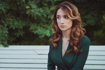 a young adult beautiful brunette woman with long curly hair is sitting on a bench in a green dress against a background of greenery.