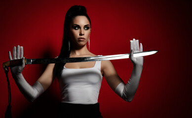 Young pretty brunette woman in white top and gloves standing and holding dagger in hands looking away over dark red background. Martial arts and beautiful women concept