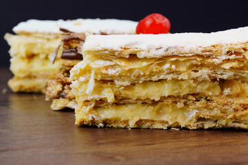 Puff pastry cakes with powdered sugar and cream and another with chocolate and a cherry on top on the wooden table. Close up. Selective focus.