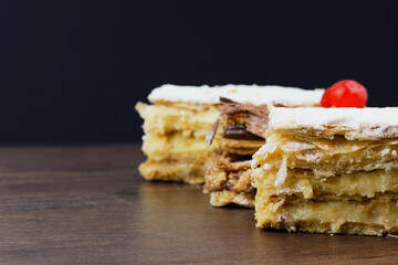 Puff pastry cakes with powdered sugar and cream and another with chocolate and a cherry on top on the wooden table. Close up and space for text. Selective focus.