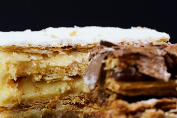 Puff pastry cakes with powdered sugar and cream and another with chocolate and a cherry on top on the wooden table. Close up. Selective focus.