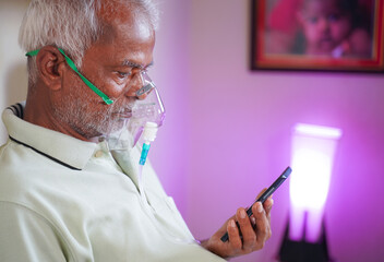 Old man with Oxygen concentrator mask reading news using mobile phone during home isolation - Concept of low oxygen level or therapy treatment from home for covid-19 coronavirus infected people