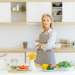 Portrait of beautiful positive mature woman in apron in kitchen looking at camera and smiling, retired grandmother standing near table with healthy food ingredients while cooking for family at home