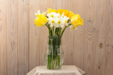 Bright bouquet of yellow and white flowers in a transparent glass jug on a wooden background. Tulips and daffodils in a glass vase.