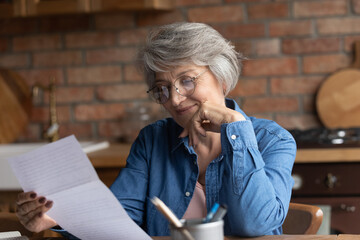 Smiling middle-aged woman in eyeglasses sit at table in kitchen reading document, receive good news...