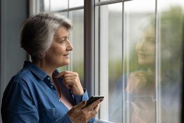 Middle aged dreamy woman holding cellphone standing near window looking outside. Grey haired older...