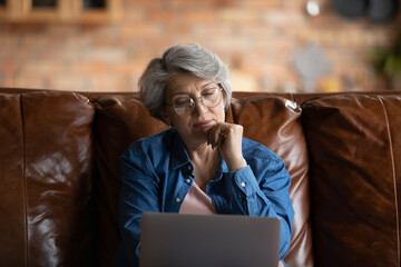 Thoughtful old woman in glasses sit on sofa with laptop, touch chin with hand having doubts looks...