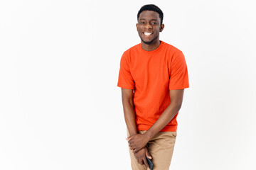 African guy in an orange T-shirt with a mobile phone and in trousers on a light background