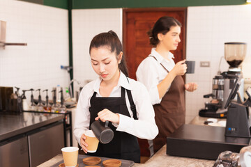 Asian barista women fill milk into takeaway hot coffee cup while caucasian barista woman holds coffee cup and talking with customer