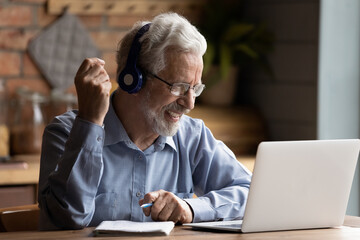 Grey haired elderly man sit indoors wear headphones looks at laptop screen holding pen gain new...