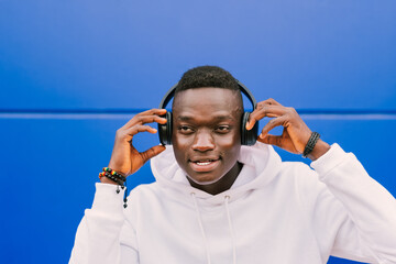 Young afro American black man listening music with wireless headphones while wearing a white sweatshirt against a blue blackground wall