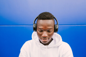 Young afro American black man listening music with wireless headphones while wearing a white sweatshirt, looking down. Sad emotion