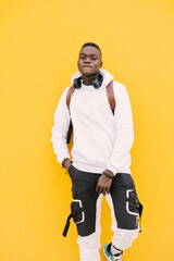 Young african American black man against a yellow wall wearing a white sweatshirt and a backpack listening music on wireless headphones while looking at camera with attitude
