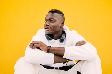 Young african American black man combing his afro hair while seated on a yellow wall background, wearing a white sweatshirt and wireless headphones. Diversity concept. BLM ethnic