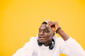 Young african American black man combing his afro hair while seated on a yellow wall background, wearing a white sweatshirt and wireless headphones. Copy space