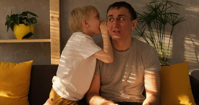 Little blond boy whispers the secret to his father in his ear sitting on the couch. Evening natural light. Slow-motion