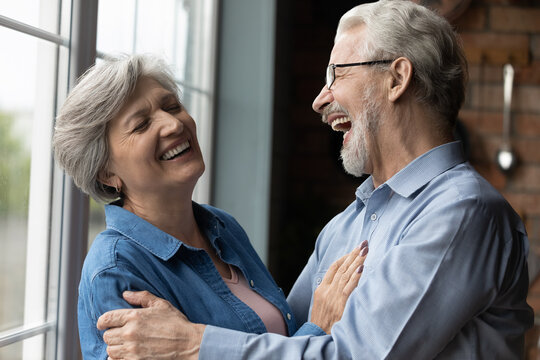 Middle-aged Untroubled Couple Happy Homeowners Hugging Laughing While Standing In Kitchen At Home. Medical Insurance Cover, Dental Clinic Services For Older Citizen, Carefree Midlife And Love Concept