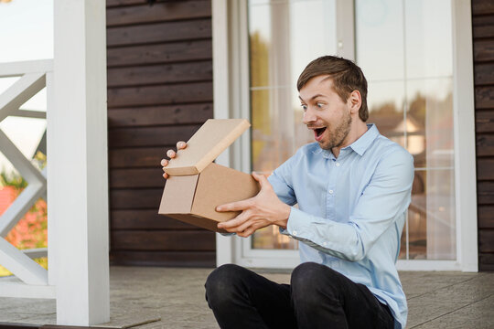 Excited young man opening box with parcel while sitting on terrace of house - Powered by Adobe