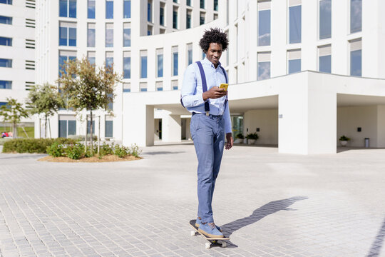 Black businessman on a skateboard looking at his smartphone outdoors.