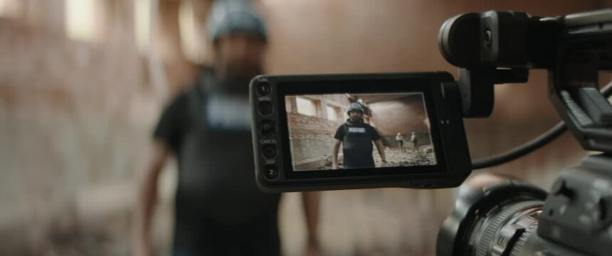 Behind the scenes of war journalist correspondent wearing bulletproof vest and helmet reporting live near destroyed building, military personnel in the background