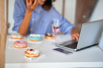 A woman is eating delicious donuts while sitting and using a laptop in a pastry shop. Pastry shop,...