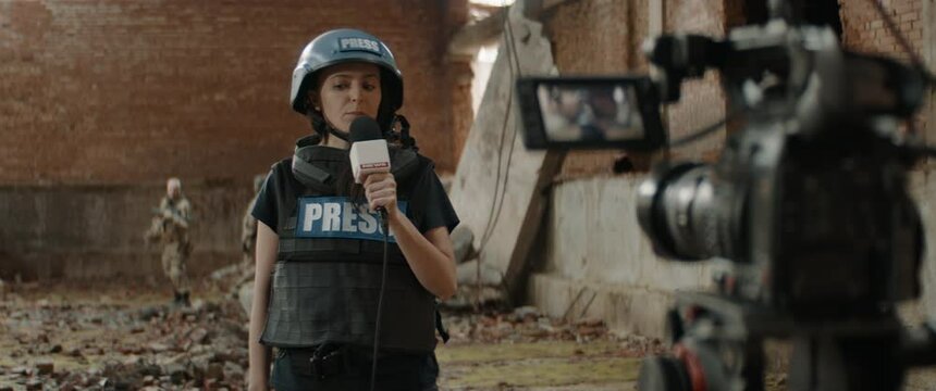 HANDHELD Behind the scenes of female war journalist correspondent wearing bulletproof vest and helmet reporting live near destroyed building, military personnel in the background