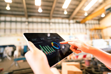Action of a working is using tablet to review storage report with blurred background of warehouse. Business management and technology concept photo. Close-up and selective focus at human's hand part.