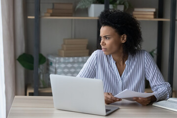 Unhappy young African American woman distracted from computer work look in distance window lack motivation. Upset displeased biracial female employee bored of monotonous job on laptop in office.