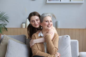 Happy daughter hugging older mother, standing behind sofa in living room, enjoying tender moment at...