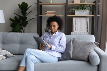 Obraz premium Happy young African American woman sit relax on couch at home use modern tablet gadget. Smiling millennial biracial female rest on sofa in living room talk on webcam video call on pad device.