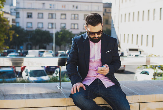 Serious male entrepreneur with sunglasses in elegant suit standing on street and surfing internet via smartphone