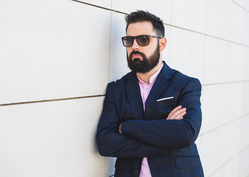 Young business man with a beard and sunglasses is dressed in a suit while leaning on the wall